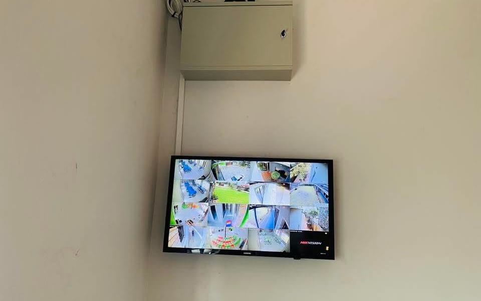 A wall-mounted screen showing sixteen camera views from a Hikvision recorder, with the recorder in a locked cabinet above it