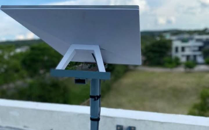 A Starlink dish on a pole mount at the edge of a flat roof, looking out over trees and houses
